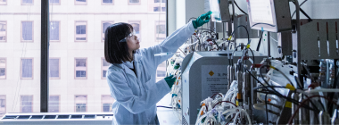 A woman in a lab coat and safety goggles reaches up towards a computer monitor.