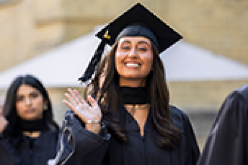 A student wearing regalia and cap smiles and waves