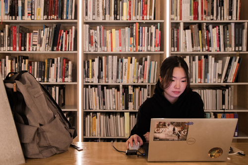 A student sits and looks a their laptop with shelves of books behind them