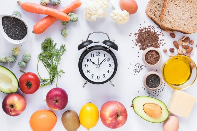 table of various fresh fruits and vegetables and grains with an analog alarm clock in the center