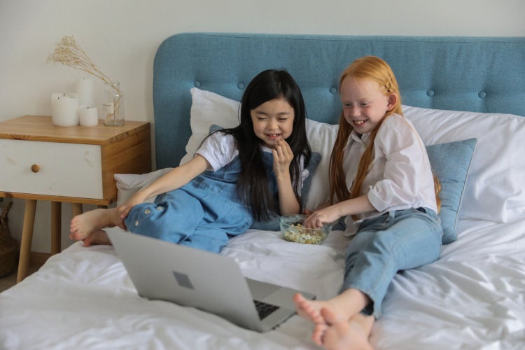 two adolescent girls watch a movie on a laptop while they sit on a bed