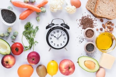 table of various fresh fruits and vegetables and grains with an analog alarm clock in the center