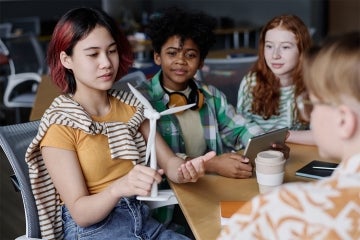 a young woman holds a model wind turbine. She is seated with other young people around a table.