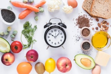 table of various fresh fruits and vegetables and grains with an analog alarm clock in the center